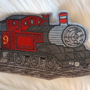 Könnte beinhalten: Ein gestickter Bügel-Patch mit einem roten und grauen Cartoon-Zug mit der Nummer 9 an der Seite. Der Zug hat einen schwarzen Schornstein und ein großes Rad hinten.