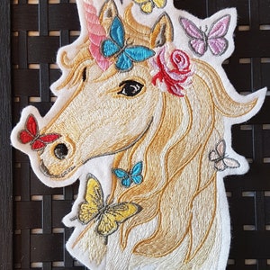 Könnte beinhalten: Gestickter Aufnäher mit einem weißen Einhorn mit einem rosafarbenen Horn und einer goldenen Mähne. Das Einhorn ist mit einer Blumenkrone und mehreren bunten Schmetterlingen verziert.
