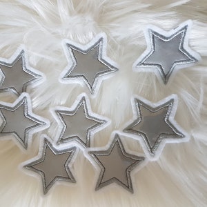 Könnte beinhalten: Acht silberne, sternförmige Bügelbilder mit weißen Umrandungen. Die Patches sind in einer Gruppe auf einer weißen, flauschigen Oberfläche angeordnet.