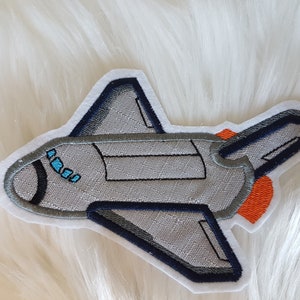 Könnte beinhalten: Ein gestickter Aufnäher einer Raumfähre in Grau und Blau mit einer orangefarbenen Flamme.