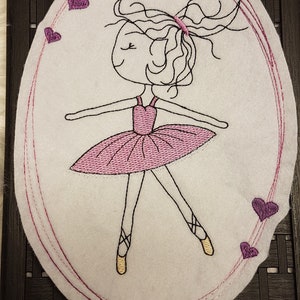Könnte beinhalten: Besticktes weißes Oval mit einem Ballerina-Design. Die Ballerina trägt einen rosa Tutu und Spitzenschuhe. Das Design ist mit schwarzem Faden umrandet, mit rosa Herzen und einem rosa Rand.
