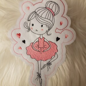 Könnte beinhalten: Gestickter Aufnäher mit einer Ballerina in einem rosa Kleid und Dutt, umrandet mit schwarzem Faden. Der weiße Aufnäher hat einen gewellten Rand mit gestickten Herzen und Pik. Das Design befindet sich auf einer flauschigen weißen Oberfläche.