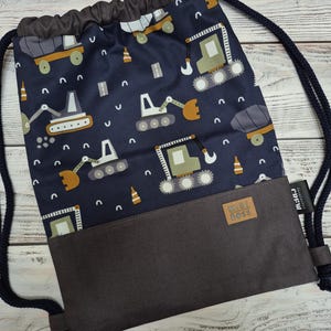 Könnte beinhalten: Dunkelblauer Kordelzugrucksack mit einem Muster aus Baumaschinen in Grün, Orange und Grau. Der untere Teil ist einfarbig dunkelgrau, mit einem kleinen braunen Etikett mit der Aufschrift "Mini Boss". Die Tasche hat marineblaue Seilriemen.