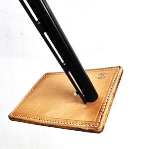 May include: A tan leather stand with a stitched edge and a small circular logo. A black metal pen is inserted into the stand.