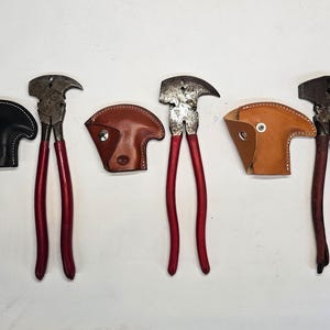 May include: A selection of vintage farrier tools, including hoof nippers with red handles and leather protective covers in black, brown, and tan. The tools are arranged on a white surface.