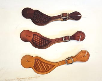 Può includere: Tre paia di cinturini per speroni in pelle marrone con disegni intrecciati. I cinturini hanno fibbie argentate e sono fissati su uno sfondo bianco.