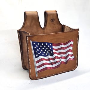 May include: A tan leather tool pouch featuring a printed graphic of a waving American flag on the front panel. The pouch has two belt loops at the top, visible white stitching along the edges, and a small circular embossed logo on the upper right.