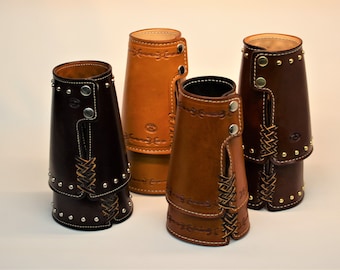 May include: Four handcrafted leather drink sleeves in varying shades of brown. Each sleeve features decorative stitching, metal studs, and a braided leather accent. The sleeves have a button closure and are designed to fit around a standard-sized beverage container.
