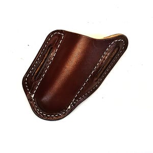 May include: A brown leather sheath with white stitching. The sheath has a curved shape and two slots on the sides. The leather has a glossy finish. The item is set against a white background.