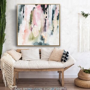 May include: Abstract painting with pink, green, and blue watercolor washes on a white canvas. The painting is framed in a gold frame and is hanging on a white wall above a white couch.