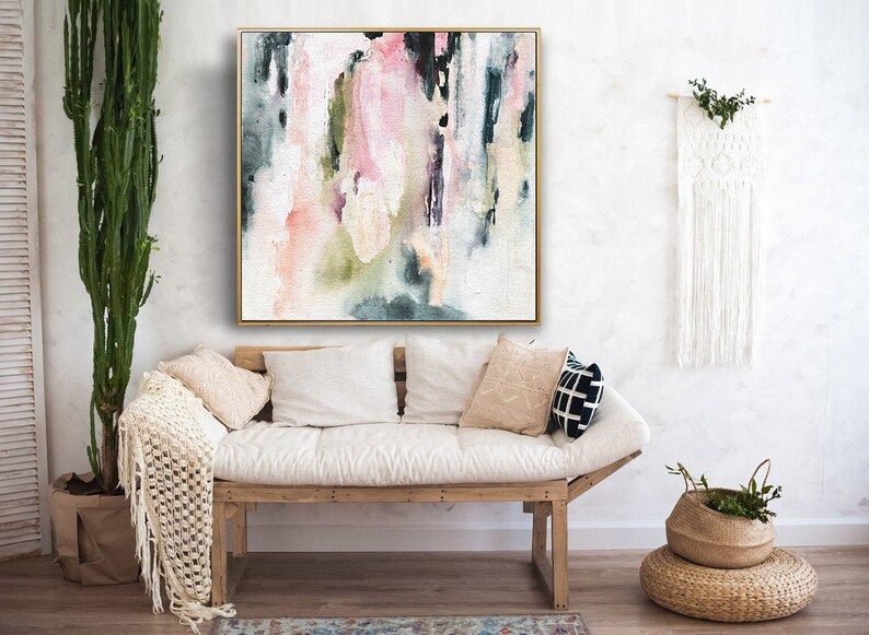 May include: Abstract painting with pink, green, and blue watercolor washes on a white canvas. The painting is framed in a gold frame and is hanging on a white wall above a white couch.