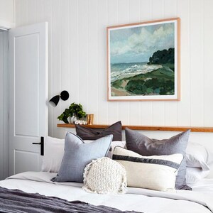 May include: A bedroom with a white bedspread and a gray throw blanket. The bed has a variety of pillows in shades of gray and blue, including a white crocheted pillow. A framed painting of a seascape hangs above the bed.