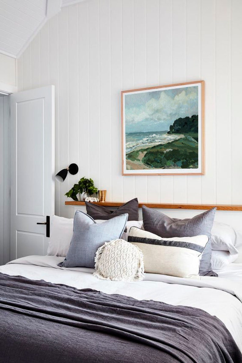 May include: A bedroom with a white bedspread and a gray throw blanket. The bed has a variety of pillows in shades of gray and blue, including a white crocheted pillow. A framed painting of a seascape hangs above the bed.