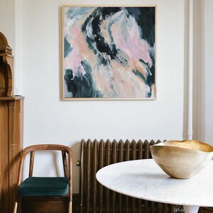 May include: A white round table with a gold bowl on top. The table is in front of a radiator and a wooden chair with a green cushion. A framed abstract painting with pink, white, and blue colors hangs on the wall behind the table.