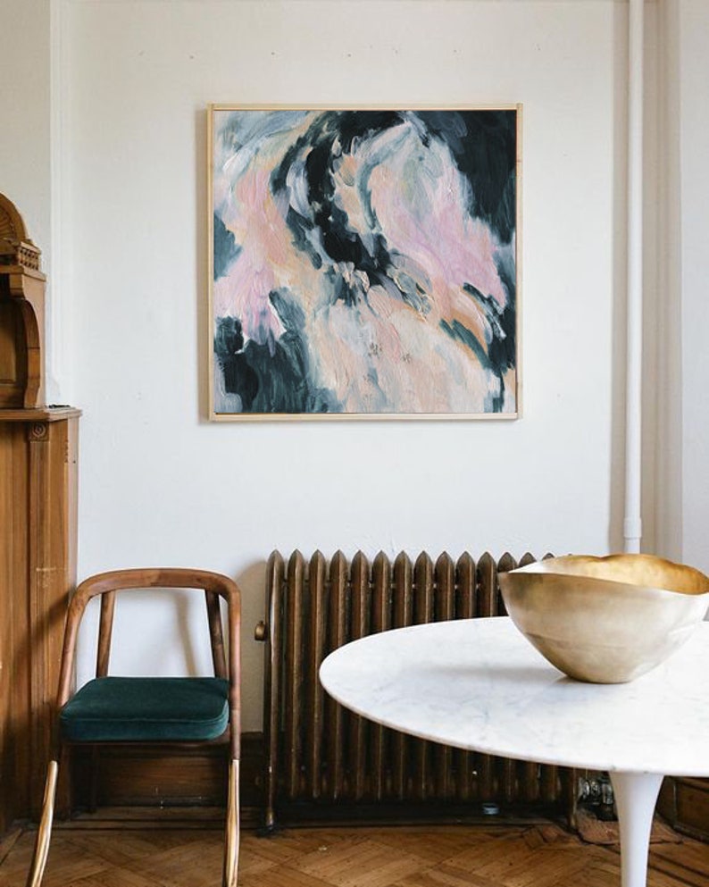 May include: A white round table with a gold bowl on top. The table is in front of a radiator and a wooden chair with a green cushion. A framed abstract painting with pink, white, and blue colors hangs on the wall behind the table.