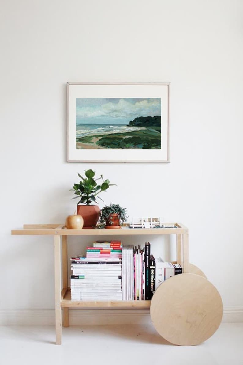 May include: A wooden rolling cart with two shelves, filled with books and magazines. The cart has a large wooden wheel on the right side. A framed painting of a seascape hangs on the wall behind the cart.