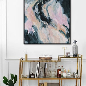 May include: A gold metal bar cart with glass shelves. The cart is filled with various bottles, glasses, and barware. A painting with abstract pink, white, and blue brushstrokes hangs on the wall above the cart.