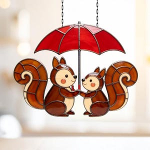 Peut inclure: Décoration en vitrail représentant deux écureuils sous un parapluie rouge. Les écureuils sont bruns et crème, avec des joues roses. Le parapluie a une armature noire et est suspendu à une chaîne. Une pièce charmante pour une fenêtre.