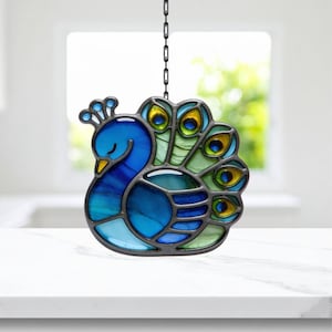 May include: A stained glass peacock suncatcher with a chain for hanging. The peacock features shades of blue, green, and yellow. The bird's body is primarily blue, with a yellow beak and a crown. The tail feathers are green and yellow.