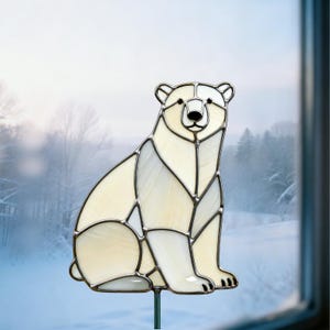 May include: A stained glass polar bear sculpture in shades of white and cream, with a black nose and eyes. The bear is sitting, with its paws and body clearly defined. The glass is outlined with a dark metal. The sculpture is mounted on a green metal rod.