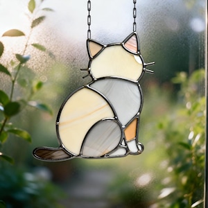 Op de afbeelding: Gebrandschilderde kattenvanger in een zittende pose. De kat heeft een mix van crème, grijze en oranje kleuren, met een donker metalen frame en ketting om op te hangen. De staart van de kat is bruin.