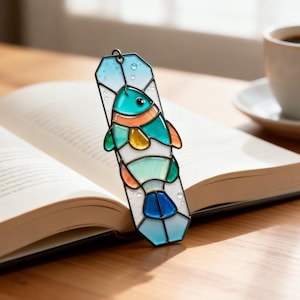 May include: A stained glass bookmark featuring a colorful fish design. The fish has teal, orange, and yellow accents, set against a white background. The bookmark is rectangular with a loop at the top, resting on an open book.