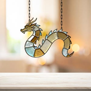 Puede incluir: Adorno de dragón de vidriera, colgado de una cadena. El dragón presenta un cuerpo segmentado en tonos marrones, dorados y azules. La cabeza y la cola son doradas, y el adorno cuelga sobre un fondo borroso. Objeto decorativo.