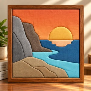 May include: A square fabric wall art piece in a wooden frame depicting a landscape with a yellow sun, orange sky, dark grey cliffs, and a blue sea. The scene features layered textile sections in shades of tan, light blue, and dark blue, creating a textured, geometric representation of a coastal sunset view.