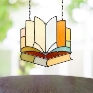 Puede incluir: Atrapasoles de vidrio manchado con forma de libro abierto, colgado de una cadena negra. El diseño del libro presenta varios colores, incluyendo naranja, amarillo, verde y rojo, creando una pieza artística.