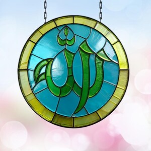 May include: A circular stained glass suncatcher featuring a central green Arabic calligraphy design set against a vibrant turquoise background. The piece is framed by a border of yellow glass segments and is suspended by two metal chains.