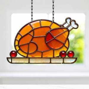 Può includere: Decorazione a vetrata per il Ringraziamento a forma di tacchino. Il tacchino è composto da pezzi di vetro arancione, giallo e rosso, con un osso bianco. È appeso a una catena nera e poggia su un vassoio bianco con tre perle di vetro rosse.