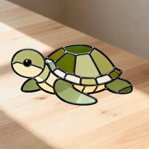 May include: A stained glass turtle figurine in shades of green, cream, and black. The turtle's shell is segmented with various shades of green, and the head and flippers are a lighter green. The figurine is placed on a light wood surface.