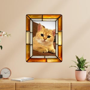 May include: A rectangular stained glass frame with a golden tabby kitten inside. The frame has a border of amber, white, and gold glass pieces. The kitten has large, round eyes and is resting on a brown surface.