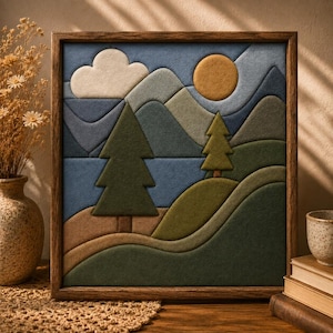 Op de afbeelding: Een vierkant wandkunstwerk in een donkerhouten lijst met een gelaagd viltlandschap. De scène toont glooiende heuvels in tinten groen en bruin, twee groenblijvende bomen, een blauw gebergte, een witte wolk en een goudgele zon.