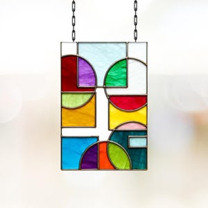 Pode incluir: Um painel retangular de vitral com um design geométrico. O painel apresenta várias formas e cores, incluindo vermelho, roxo, verde, amarelo e azul. É suspenso por uma corrente preta, adicionando um toque decorativo à obra de arte.