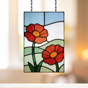 Puede incluir: Panel de vidriera rectangular con dos amapolas rojas con centros amarillos, tallos y hojas verdes. El fondo incluye un cielo azul y colinas verdes. El panel está enmarcado en negro y colgado de una cadena.