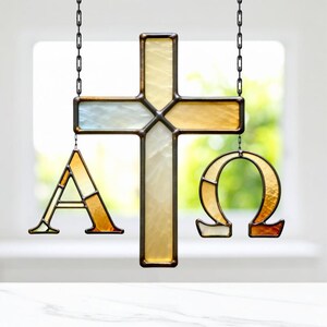 May include: Stained glass cross with the Greek letters Alpha and Omega hanging below. The cross and letters are made of amber, cream, and blue-tinted glass, framed with a dark metal. The piece hangs from a chain.