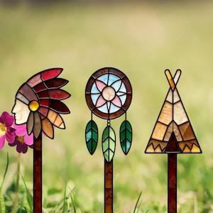 Puede incluir: Tres estacas de jardín de vidrio tintado que muestran el perfil de una persona con un tocado de plumas, un atrapasueños circular con tres hojas colgantes y un tipi triangular, montados sobre varillas verticales de color marrón oscuro.