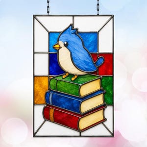 Puede incluir: Un panel de vidriera rectangular que presenta un pájaro azul con el pecho color crema posado sobre una pila de tres libros. Los libros son verdes, azules y rojos con páginas color crema. El fondo consta de paneles de vidrio geométricos en blanco, azul, rojo, verde y amarillo.