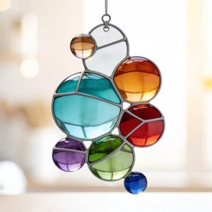 May include: A colorful stained glass suncatcher with a cluster of round, jewel-toned glass pieces. The colors include blue, green, purple, red, orange, and clear. The glass is held together by a silver-colored metal frame, and it hangs from a small chain.