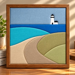 May include: A square wall art piece featuring a fabric-layered coastal landscape in a wooden frame. The scene depicts a white lighthouse against a light blue sky, a dark blue ocean, a turquoise wave, a beige sandy path, and green grassy hills.
