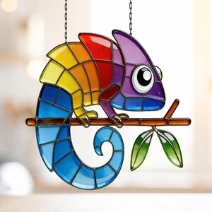 Puede incluir: Un atrapasoles de cristal con un camaleón colorido. El camaleón está posado en una rama marrón con hojas verdes. El cuerpo es un arcoíris de colores, incluyendo rojo, naranja, amarillo, azul y morado. Cuelga de una cadena.
