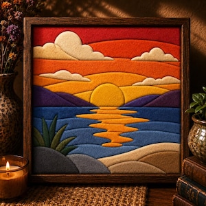 Puede incluir: Una pieza de arte mural cuadrada y enmarcada que presenta un paisaje de fieltro en capas de una puesta de sol sobre el agua. La escena muestra un sol amarillo brillante, un cielo naranja y rojo con nubes crema, montañas moradas y una playa.