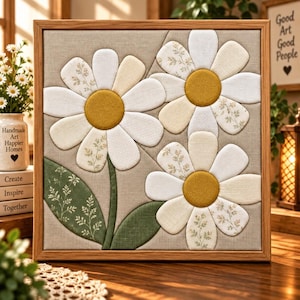 May include: A square wall art piece featuring three fabric daisies with white and patterned petals and mustard yellow centres. The flowers are set against a light beige linen background with green fabric leaves. The artwork is encased in a wooden frame.