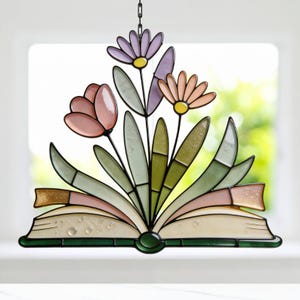 May include: Stained glass window hanging featuring an open book with colorful flowers. The flowers include pink tulips, lavender and peach daisies, and green leaves. The book has a green spine and gold-colored pages.