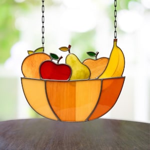 May include: Stained glass fruit bowl with a variety of fruits, including an orange, red apple, pear, and banana. The bowl is orange and yellow, and the fruits have green leaves. The piece hangs from a black chain.