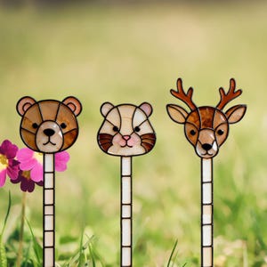 May include: Three stained glass garden stakes featuring animal heads: a brown bear, a light tan hamster, and a brown deer with antlers. Each animal head is mounted on a long, thin, white-tiled stake with black grout lines between the glass pieces.