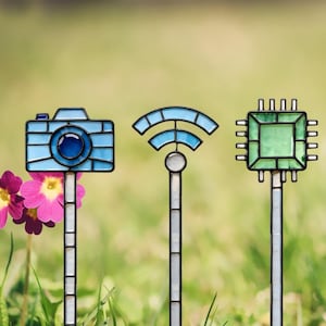 May include: Three stained glass garden stakes featuring a blue camera, a blue Wi-Fi symbol, and a green computer processor chip. Each piece is constructed with dark metal framing and mounted on a vertical stake, showcasing geometric glass segments.