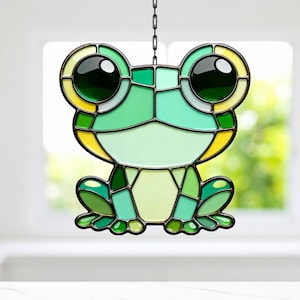 Puede incluir: Atrapasoles de rana de vidrieras con una cadena para colgar. La rana presenta una variedad de tonos verdes, acentos amarillos y ojos negros grandes. El diseño recuerda a un personaje de dibujos animados.