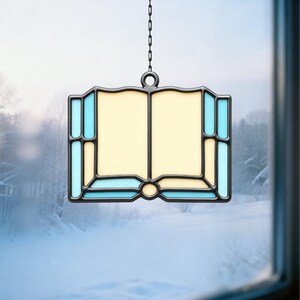 May include: A stained glass window panel in the shape of an open book, hanging from a chain. The book's pages are a soft yellow, framed by a black border with light blue accents. The background shows a snowy winter scene.